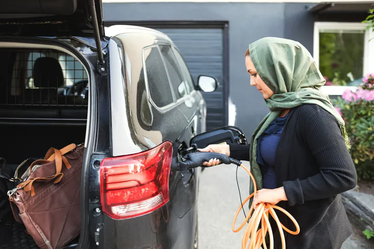 Side view of woman charging electric car with open trunk while standing outside house