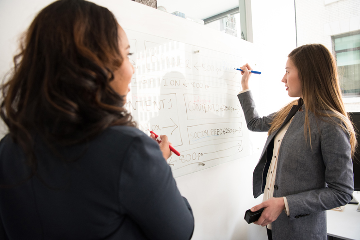 Two women standing in front of a rectangular whiteboard