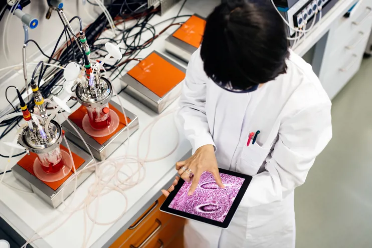 A female Asian scientist is doing research in a laboratory. She's working on a tablet computer next to a desk with flasks filled with substances and other scientific equipment.
